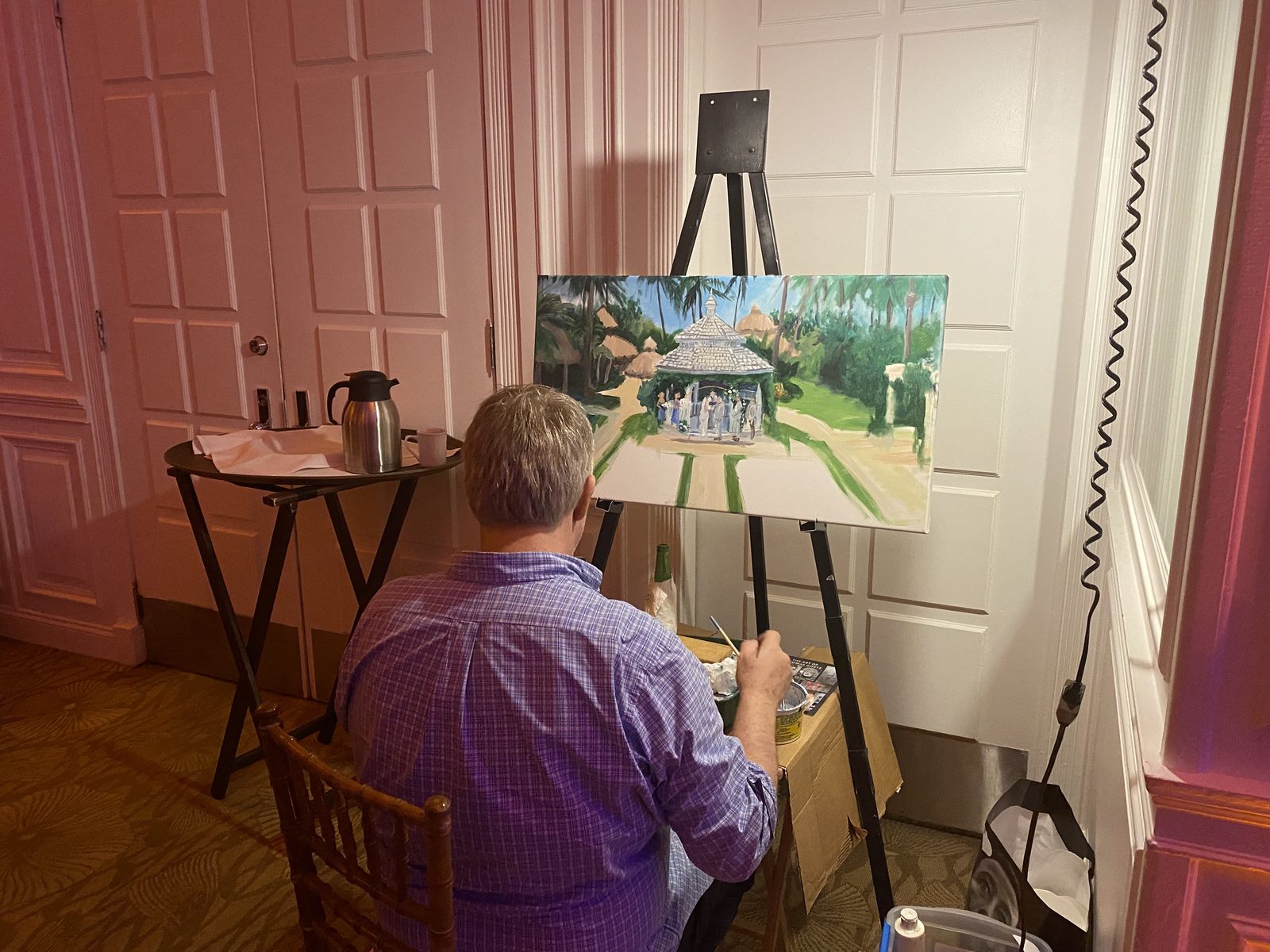 Tom Goetz seated at an easel in a wedding reception room, painting the couple's outdoor ceremony under a white gazebo.